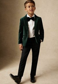 Blazer en velours vert avec un pantalon noir et une chemise blanche, agrémenté d'un nœud papillon noir et de chaussures habillées noires brillantes. Design simple et élégant.