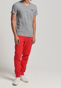 T-shirt grigio con scollatura a V, maniche corte, abbinato a pantaloni della tuta rossi con un logo nero. Sneakers bianche completano l'outfit.