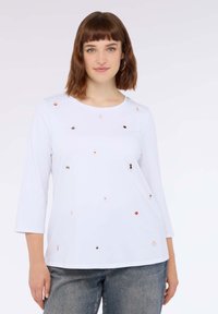 Top bianco a maniche lunghe con scollo rotondo, decorato con paillettes colorate di varie forme. Abbinato a jeans in denim grigi.