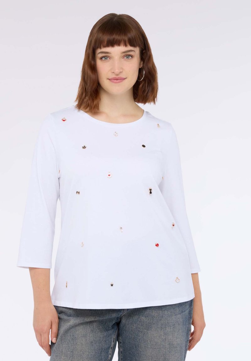Top bianco a maniche lunghe con scollo rotondo, decorato con paillettes colorate di varie forme. Abbinato a jeans in denim grigi.