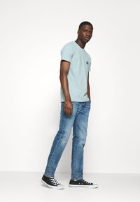 Maglietta di cotone azzurro chiaro con maniche corte, logo nero sul petto, abbinata a jeans in denim blu medio e sneakers nere alte.