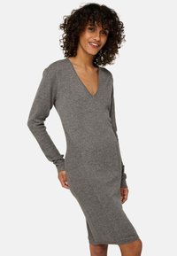 Femme avec des cheveux bouclés portant une robe en tricot grise à manches longues, arrivant au genou et à col en V, souriant et posant devant un fond uni.