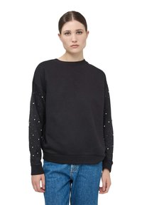 Czarna bluza z długimi rękawami, ozdobiona złotymi ćwiekami na rękawach, okrągłym dekoltem i luźnym krojem, zestawiona z niebieskimi dżinsami.