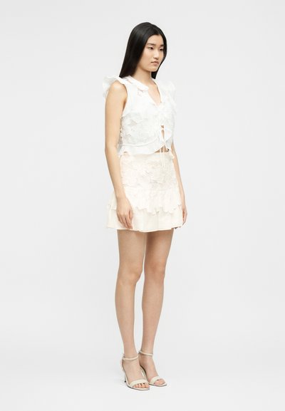 Νεαρή γυναίκα που φοράει λευκό κορυφαίο χωρίς μανίκια από δαντέλα και ταιριαστό μίνι φούστα με βολάν, καθώς και τακούνια με λουράκι στον αστράγαλο, στέκεται μπροστά σε μια απλή φόντο.