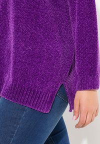 Lila texturerad tröja med ribbade muddar och nederkant, med sidfickor, tillsammans med blå jeans. Närbild av materialet och detaljerna.