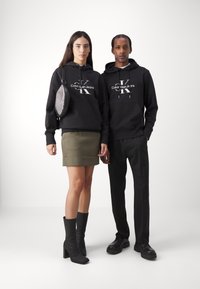 Crne dukserice s bijelim logotipom "Calvin Klein Jeans", u paru s maslinasto zelenom suknjom i crnim čizmama s visokom petom, u minimalnoj pozadini.