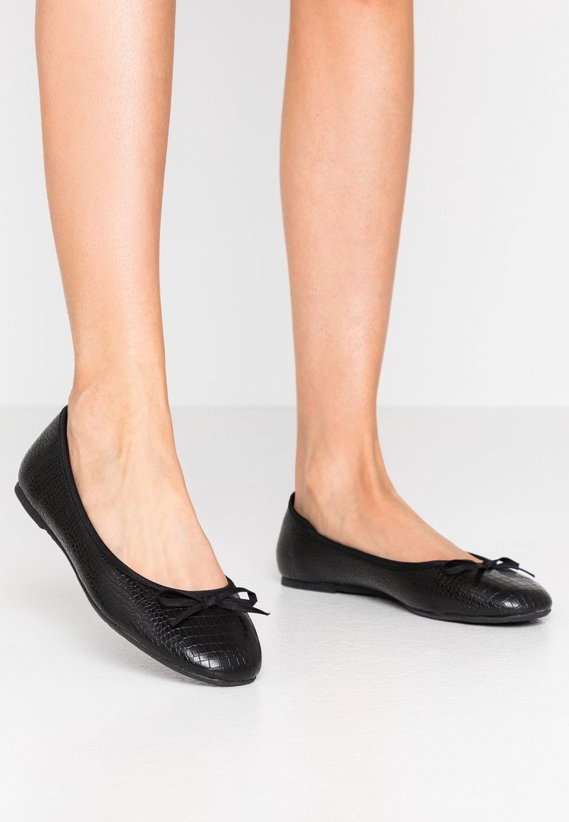 Pieds chaussés de ballerines noires texturées avec petits nœuds, posés sur un sol blanc devant un fond neutre.