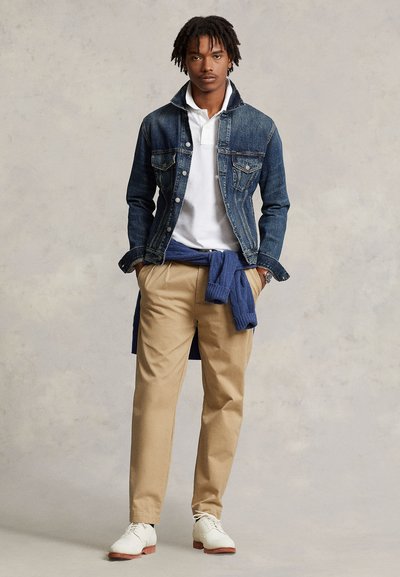 Jeune homme debout portant une veste en jean sur un polo blanc, un pantalon kaki, des chaussures blanches, avec un pull bleu noué autour de la taille.