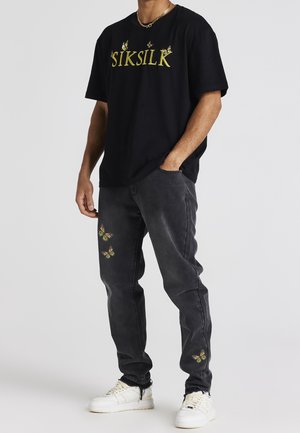 Mężczyzna w czarnej koszulce SikSilk z żółtymi motywami motyli i ciemnych dżinsach z nadrukiem motyli w pobliżu kieszeni, białe trampki i złoty łańcuszek.