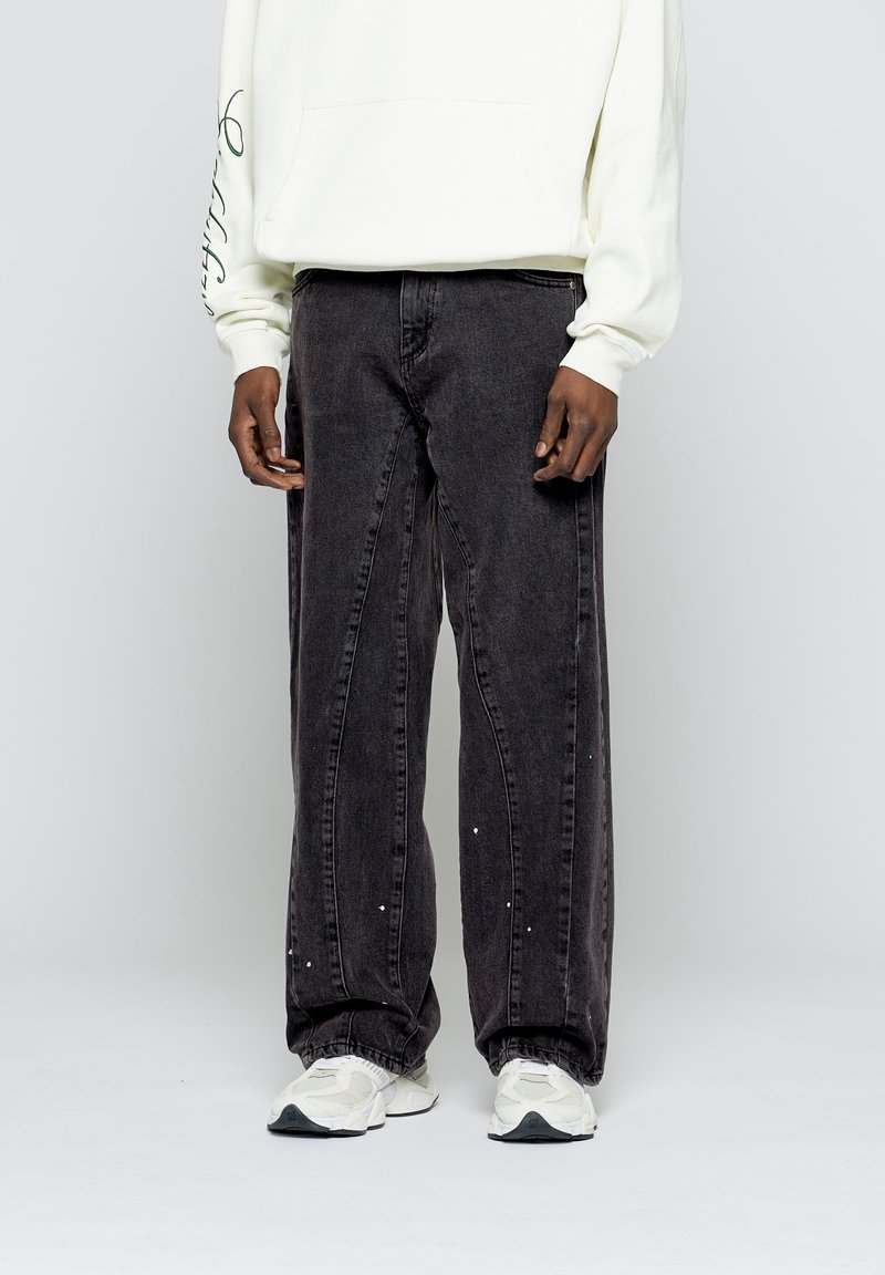Homme portant un jean noir ample avec des éclaboussures de peinture blanche, des baskets blanches et un sweat blanc avec un texte sur une manche.