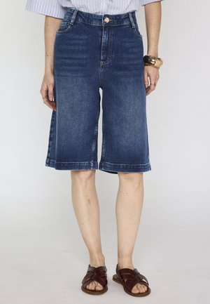 Femme portant un short en denim bleu foncé à hauteur des genoux, des sandales marron et un ourlet de chemise rayée visible, debout devant un fond uni.