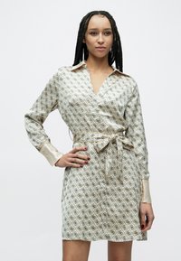 Vzory šiat na zavinovanie v krémovej farbe s čiernymi geometrickými tvarmi. Má dlhé rukávy, opasok v páse a saténový golier a manžety.