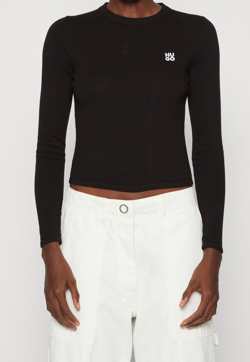 Personne portant un haut noir cintré à manches longues avec le logo "HUGO" et un pantalon blanc taille haute avec une fermeture bouton.