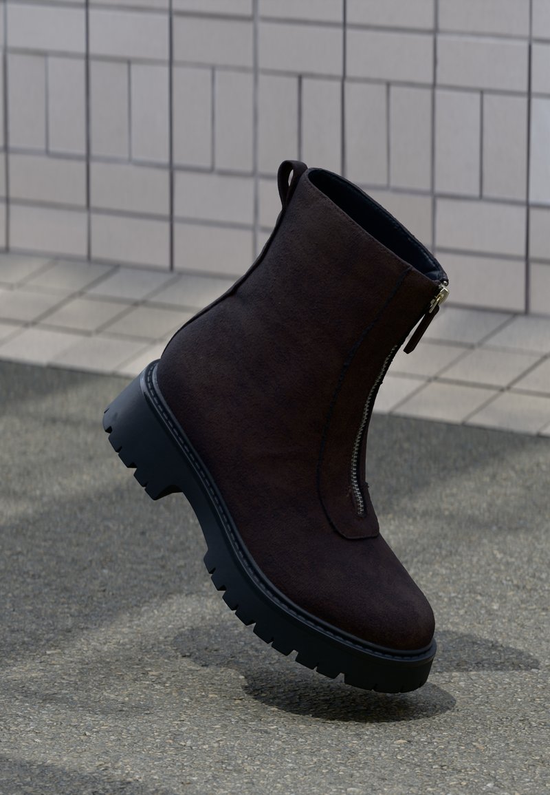 Brown suede ankle boot with a textured surface, side zipper, and rugged black rubber sole, featuring a slightly elevated heel and pull tab.