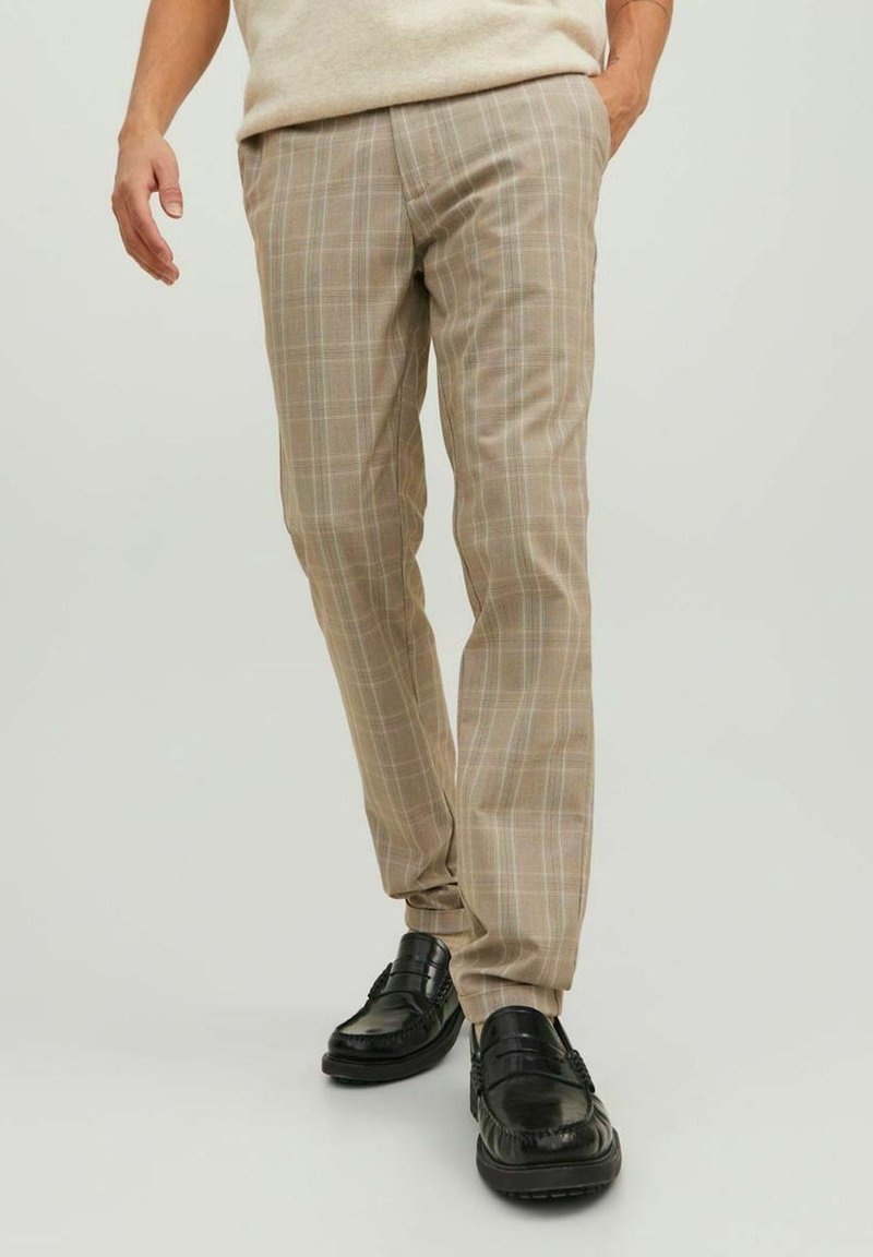 Homme portant un pantalon slim beige à carreaux et des mocassins en cuir noir, debout avec une main dans la poche sur un fond uni.