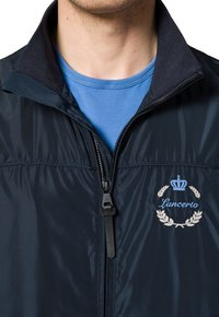 Granatowa kurtka zapinana na zamek z teksturowanym kołnierzem i haftowanym logo z koroną i laurowym wieńcem. Pod nią widać jasnoniebieską koszulę.