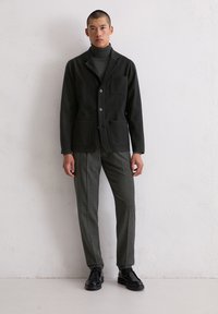 Dunkler Wollblazer mit Schalkragen, zwei Fronttaschen, Knopfverschluss, kombiniert mit grauer, maßgeschneiderter Hose und schwarzen Schnürschuhen.