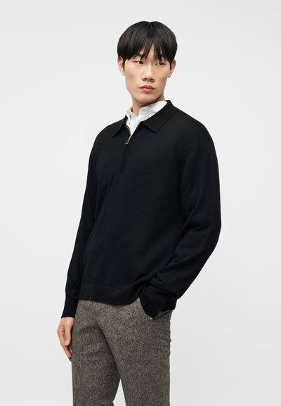 Νεαρός άνδρας με μαύρα μαλλιά, ντυμένος με μαύρο πουλόβερ με φερμουάρ πάνω από λευκό πουκάμισο και γκρι υφασμένα παντελόνια, στέκεται με το ένα χέρι στην τσέπη.