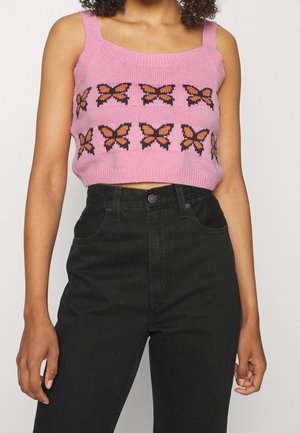 Femme portant un crop top en maille rose avec des motifs papillons orange et noir, associée à un jean noir taille haute.