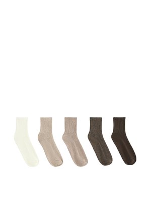 Paire de pieds portant des chaussettes chevilles côtelées beige sur un fond clair.