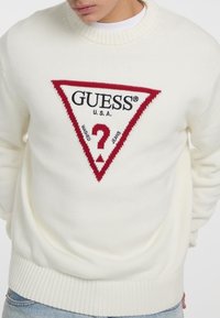 Cremefarvet strikket sweater med et rødt trekantet logo med "GUESS" og et spørgsmålstegn, ribbet halsudskæring og manchetter.