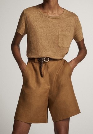 Person wearing a brown short-sleeve shirt with pocket, high-waist brown shorts, and a braided belt with hands in pockets.