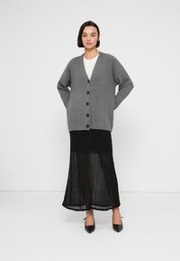 Γκρι πλεκτή ζακέτα με V-λαιμόκοψη και πέντε μαύρα κουμπιά, συνδυασμένη με μαύρη πλισέ φούστα και μαύρες μυτερές γόβες.