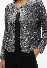 Szara marynarka z cekinami, z okrągłym dekoltem i zapięciem z przodu, o fakturowanej powierzchni i długich rękawach. Noszona na czarnej bluzce.