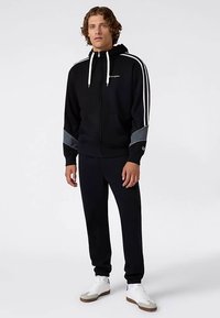 Sudadera con capucha negra con cremallera, con detalles en blanco y gris, con rayas laterales, capucha, bolsillos frontales, combinada con pantalones negros y zapatillas blancas.