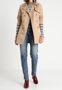 Trench-coat beige avec un design croisé et de grands revers, associé à un jean bleu et des bottines bordeaux, un haut rayé à manches longues visible.