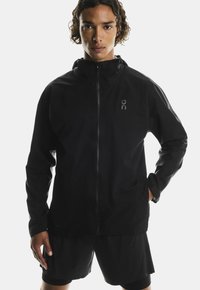 Giacca nera impermeabile con zip integrale, colletto alto e logo sul petto. Texture liscia e design aderente, abbinata a pantaloni corti neri.