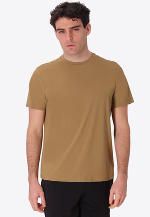 Giovane uomo con capelli ricci scuri che indossa una maglietta marrone semplice a maniche corte e pantaloni neri, in piedi su uno sfondo bianco.