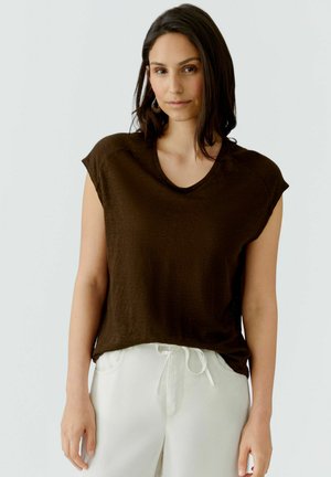 Vrouw met donker schouderlang haar, gekleed in een donkerbruin shirt met korte mouwen en witte broek met trekkoord, staand tegen een effen achtergrond.