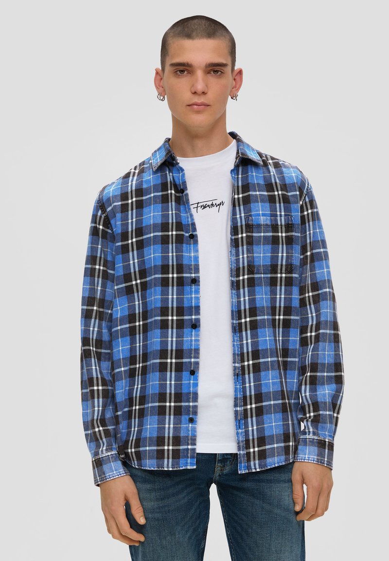 Blauwe en zwarte geruite flanellen overhemd met lange mouwen, knoopsluiting en een voorkantzak, gelaagd over een wit graphic t-shirt.