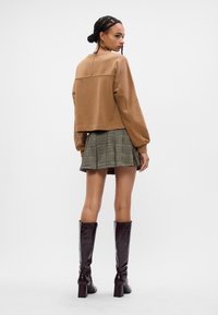 Fată cu părul împletit, îmbrăcată cu un top lung de culoare maronie, o fustă mini cu carouri și cizme înalte, de culoare maronie închisă, întorcându-se cu spatele, pe un fundal alb.