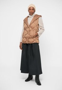 Rjav podložen brezrokavnik s kapuco, ki ima velike sprednje žepke, v kombinaciji z svetlo sivo rolko in črno tekočo krilo. Črne gležnjarje.