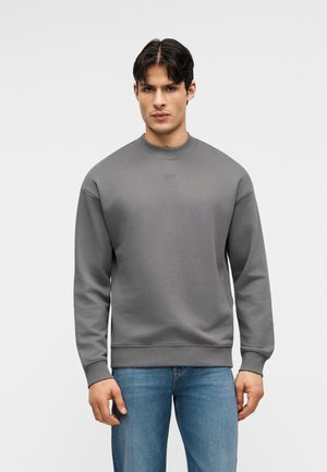 Moški, oblečen v preprosto sivo jopico z dolgimi rokavi in modre kavbojke, stoji obrnjen naprej na svetlo sivi podlagi.