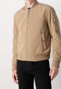 Tan bomberjacka med glänsande finish, ribbade muddar och nederkant, framdragkedja, sidofickor och en ficka med dragkedja på ärmen med logotyp.