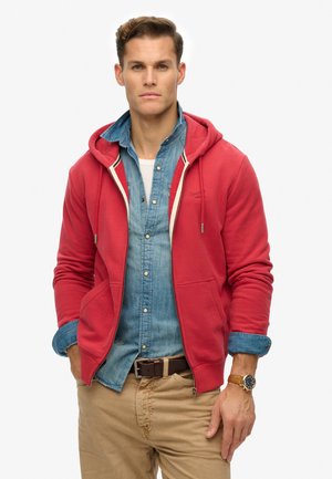 Sudadera roja con cremallera y capucha ajustable, combinada con una camisa de mezclilla. Destacan los bolsillos frontales y los puños en contraste.