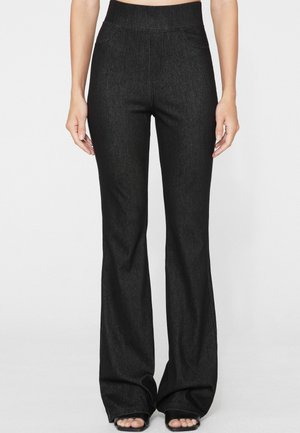 Donna che indossa pantaloni neri a vita alta e svasati abbinati a sandali neri open toe, in piedi davanti a uno sfondo chiaro e uniforme.