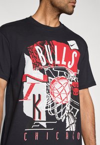 Czarny T-shirt z grafiką przedstawiającą napis „BULLS”, motywy koszykówki oraz tekst „CHICAGO” w czerwonych i białych akcentach. Obecne logo Nike.
