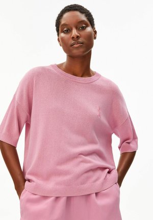 Vrouw met kort krullend haar die een losse roze gebreide top en bijpassende roze broek draagt, staand met handen in de zakken tegen een witte achtergrond.