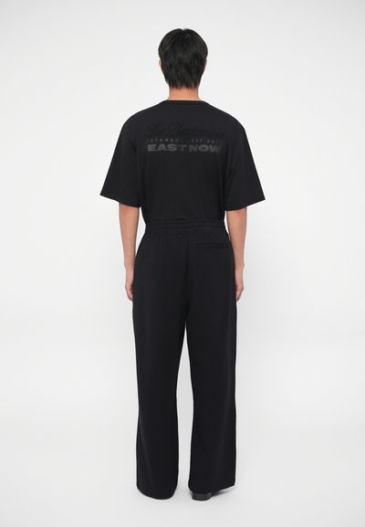 Camiseta negra con cuello redondo y mangas cortas, que presenta una impresión en relieve en la parte posterior. Combinada con pantalones negros de corte holgado.