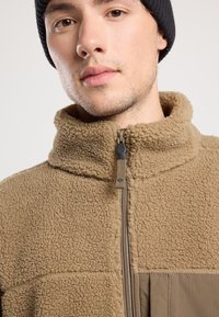 Giacca in pile marrone con colletto alto, chiusura zip e dettagli in tessuto. Superficie testurizzata, materiale morbido, presenta una tasca con zip.