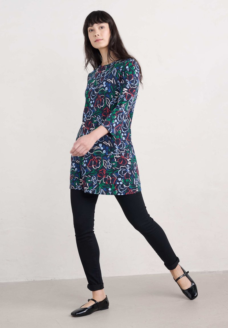Blusentunika mit floralem Muster in Marineblau und roten, grünen sowie blauen Akzenten. Lange Ärmel, gerade geschnitten, kombiniert mit schwarzen Slim Jeans und schwarzen Ballerinas.