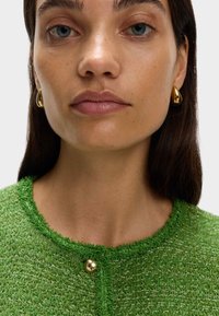 Cardigan in maglia verde con texture e bottone dorato, abbinato a orecchini a goccia metallici dorati. Pelle liscia e trucco naturale sono visibili.