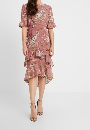 Kvinde iført en lyserød blomsterprintet high-low kjole med flæser og korte ærmer, kombineret med ankelsandaler med slangeprint.
