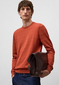 Giovane uomo in un maglione testurizzato color ruggine e jeans blu che tiene una borsa a tracolla in pelle marrone scuro su uno sfondo semplice.