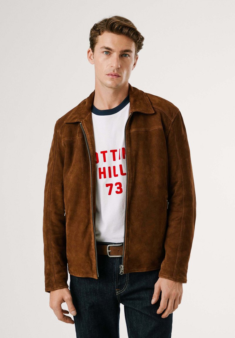 Giacca in suede marrone con collo a camicia, chiusura con zip e due tasche laterali, indossata sopra una maglietta bianca con testo rosso e jeans scuri.