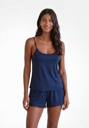 Frau lächelt, trägt ein dunkelblaues Spaghettiträger-Top und passende Shorts mit transparentem Besatz, steht vor einem schlichten weißen Hintergrund.